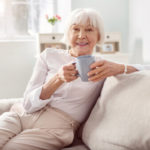 An elderly woman sits at home with a cup of coffee on her couch