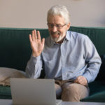 An older man waves at family members during a virtual call on his laptop