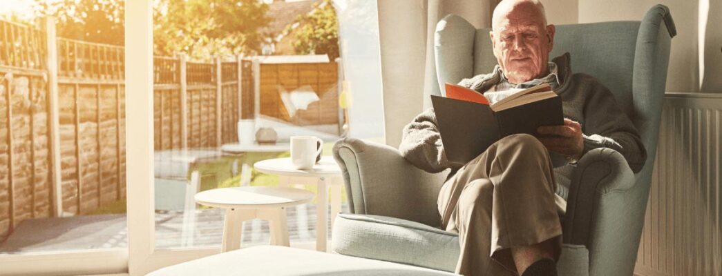 An older man sits in an armchair reading a book.