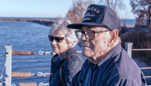 An older male veteran with his wife by the sea with a ship in the background
