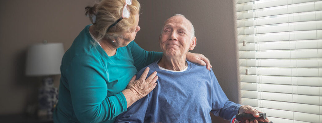 n elderly man sitting by a window, holding a cane, and smiling warmly at an elderly woman who is standing beside him, gently resting her hand on his shoulder.