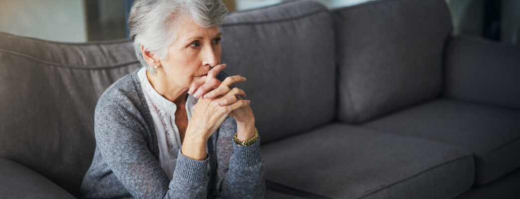 Elderly woman sitting on a couch, deep in thought, with her hands clasped in front of her face