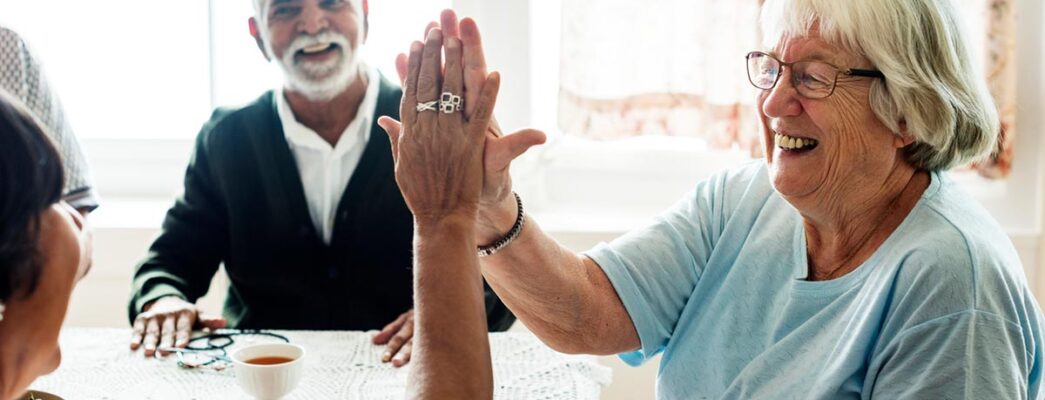 An older woman high fives another older woman sitting at a kitchen table