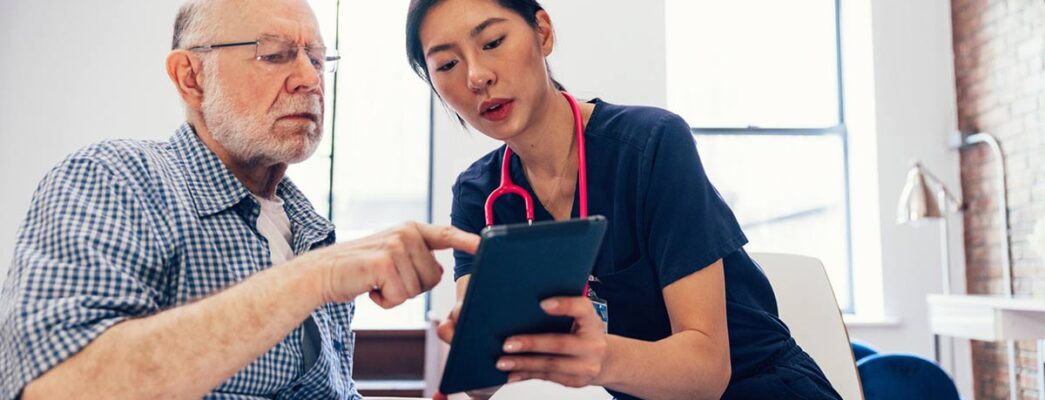 A woman with a stethoscope views a tablet with an older man who may be a patient.