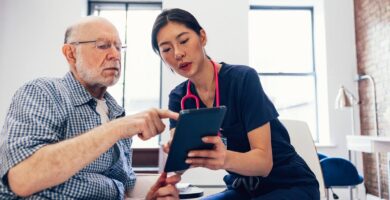 A woman with a stethoscope views a tablet with an older man who may be a patient.