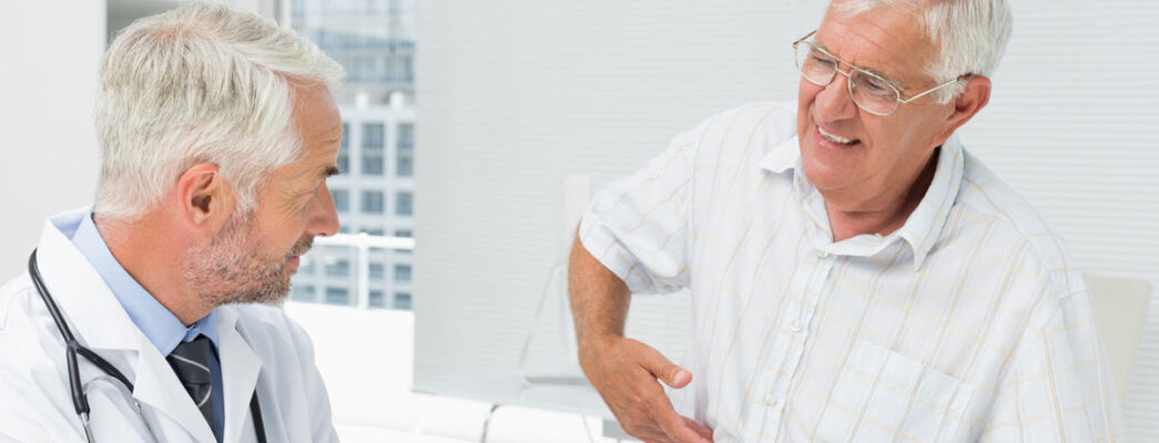 An older man in a striped shirt sits in a medical office, pointing to his side and appearing to be in discomfort, while speaking with a male doctor who is listening attentively
