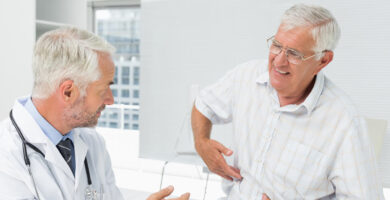 An older man in a striped shirt sits in a medical office, pointing to his side and appearing to be in discomfort, while speaking with a male doctor who is listening attentively