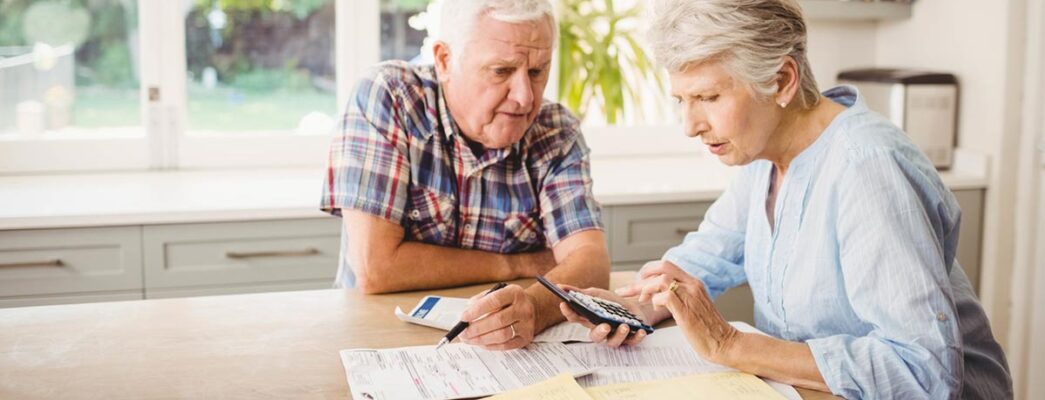 An older couple sits at their kitchen table reviewing bills with a calculator