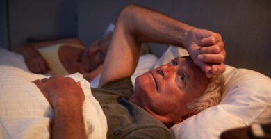 Older man lying awake in bed at night, looking worried while his partner sleeps beside him