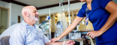 A cancer patient sits in a treatment chair receiving IV therapy while a nurse gently touches his arm in a supportive gesture