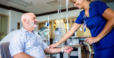 A cancer patient sits in a treatment chair receiving IV therapy while a nurse gently touches his arm in a supportive gesture