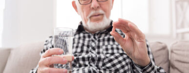 An older man holds a glass of water in one hand and a small pill in the other