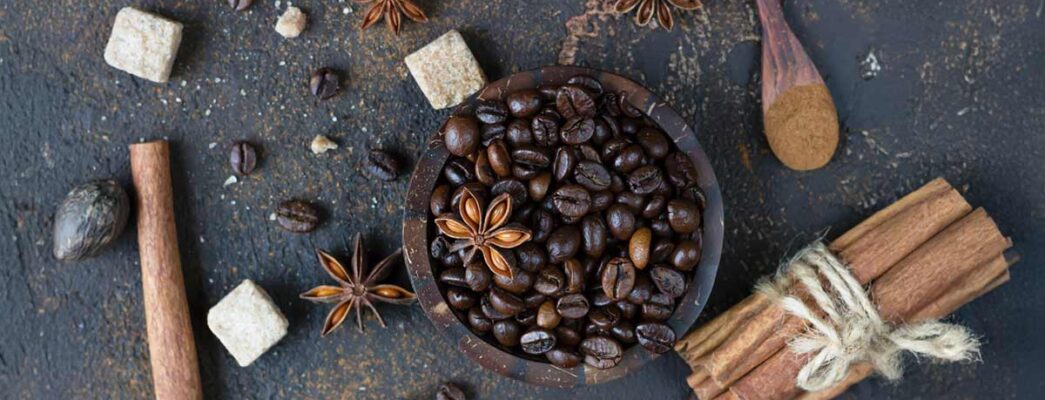 Dark roasted coffee beans in a wooden bowl surrounded by star anise, cinnamon sticks, brown sugar cubes, nutmeg, and a wooden spoon with ground cinnamon on a textured dark background.