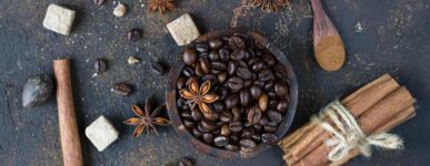 Dark roasted coffee beans in a wooden bowl surrounded by star anise, cinnamon sticks, brown sugar cubes, nutmeg, and a wooden spoon with ground cinnamon on a textured dark background.