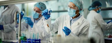 Researchers in protective lab gear, including masks, gloves, gowns, hairnets, and goggles, work in a laboratory setting.
