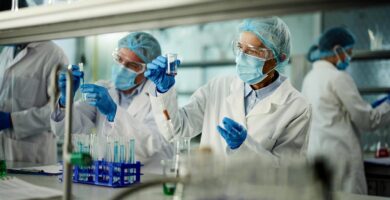 Researchers in protective lab gear, including masks, gloves, gowns, hairnets, and goggles, work in a laboratory setting.