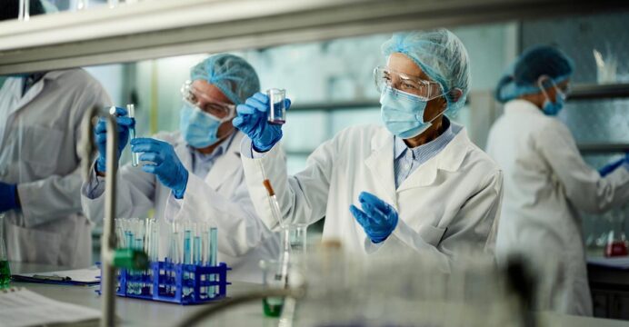 Researchers in protective lab gear, including masks, gloves, gowns, hairnets, and goggles, work in a laboratory setting.