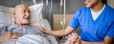 Nurse in scrubs smiling and holding the hand of an older male patient lying in a hospital bed during treatment