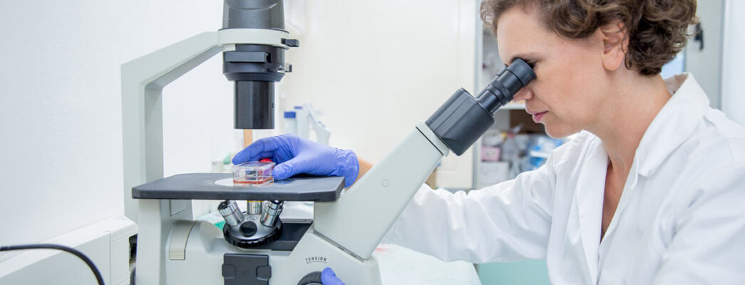 A woman wearing a lab coat examines a sample under a microscope